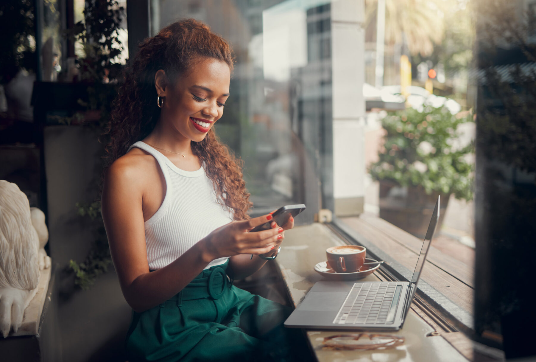 A woman in a white sleeveless top sits by a window at a cafe, smiling at her phone. A laptop and a cup of coffee are on the table in front of her. Sunlight and outdoor scenery are visible through the window.
